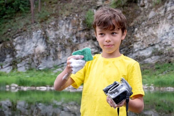Sarı bir tişört bir çocuk bir objektif olmadan elinde bir DSLR kamera tutar ve içine su ekler. Doğada bir dağ nehirde çılgın temizlik fotoğraf ekipmanları.