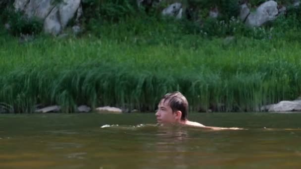 Ο έφηβος κολυμπάει δεξιά αριστερά κατά μήκος του ποταμού πέρα από τις καλαμιές. Καλοκαιρινή διασκέδαση. Διακοπές και ταξίδια. — Αρχείο Βίντεο