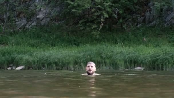 Un tipo con barba se sumerge y salta del río contra el telón de fondo de las montañas. Diversión de verano en el agua en la naturaleza. Bañarse, descansar y viajar . — Vídeo de stock