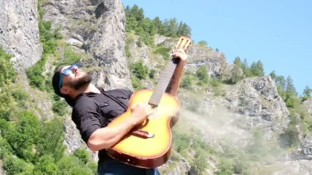 검은 셔츠와 선글라스를 입은 수염난 남자가 연기가 자욱한 어쿠스틱 기타를 연주하는 척합니다. 산에서 자연 배경에 뮤직 비디오에 대한 이상한 재미있는 비디오. — 비디오