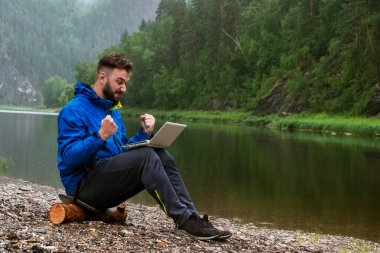 Yağmurda başarılı elektronik işlem. Nehir kıyısında bir laptop üzerinde çalışan sakallı bir adam. Su koruyucular ile dağlarda kötü havalarda Freelancer gezgin veya turist IT ekipmanları.