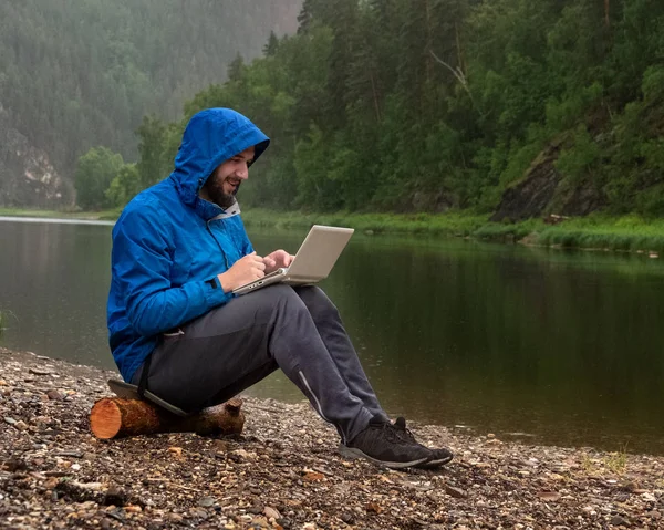 Um cara barbudo satisfeito em um casaco azul com um capuz se senta na margem de um rio na chuva com um laptop em seu colo. Freelancer nas montanhas com moderno equipamento de TI à prova d 'água em mau tempo  . — Fotografia de Stock