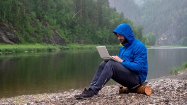 Bir Hood ile mavi ceket bir adam nehir tarafından yağmurda bir günlük otururken beyaz bir laptop üzerinde çalışıyor. Programcı veya Hacker dağlarda. Konsept su geçirmez bilgisayar donanımları.