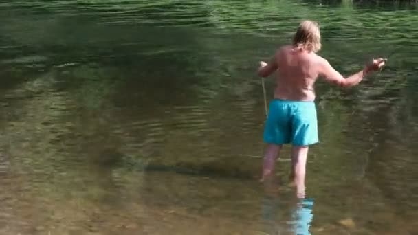山の川で年配のフライフィッシャー サンタクロースの夏休みのコンセプト 裸の背中を持つ青いショートパンツのトップレスの男は 水の中で膝深く立って餌を投げる スポーツまたは趣味 — ストック動画