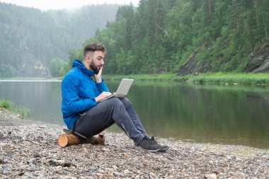 Sakallı bir genç adam su geçirmez bir bilgisayarın ekranında sürpriz görünüyor. Mavi ceketli bir adam yağmurda dizlerinin üzerinde dizüstü bilgisayarla dağ nehrinin yakınındaki bir kütükte oturuyor. Doğada işadamı.