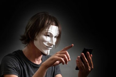 A man with long hair in a hacker or anonymous mask in a T-shirt is holding a mobile phone in his hands. The concept of an Internet hacking site, personal data or secret of cybercriminals. Gray dark. clipart