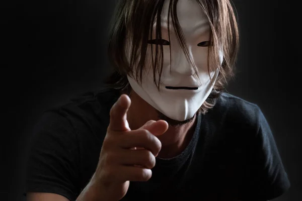 A man with long hair in a hacker or anonymous mask in a T-shirt stands with his finger forward. Concept of Internet hacking or the mystery of cybercriminals. Gray dark background. — Stock Photo, Image