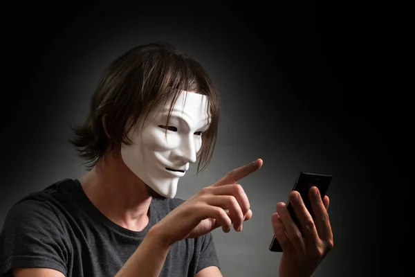 Muž s dlouhými vlasy v hackeru nebo anonymní masce v tričku drží v rukou mobilní telefon. Koncept internetové stránky hackingu, osobních dat nebo tajemství počítačových zločinců. Tmavě šedá. — Stock fotografie