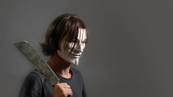 Человек в хакере или анонимной маске держит мачете на плече. Парень-злодей с длинными волосами, одетый в футболку, стоит в профиле на сером фоне. Принято. Хэллоуин . — стоковое фото