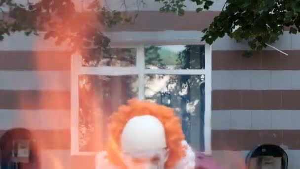 En cosplayer med smink och en kostym som en skrämmande clown Pennywise visas från elden i bakgrunden av en byggnad med telefonautomater och fönster. Halloween firande eller alla helgons dag maskerad. — Stockvideo