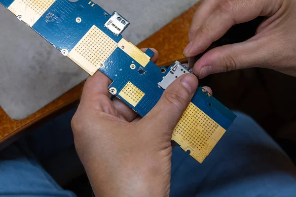 Un técnico de reparación electrónica limpia los contactos de la placa de circuito con una cuchilla afilada. Un tablero electrónico de algún artilugio en manos de un hombre . — Foto de Stock