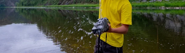 Камера в детской руке, на которую льется поток воды на фоне реки или озера с зелеными растениями. Странный способ мыть оборудование или чистить матрицу. Панорамный снимок — стоковое фото