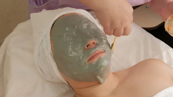 Zabieg odmodowania kosmetycznego. Szara maska alginian na twarzy azjatyckiej kobiety. Ręce kosmetyczki umieścić dużo na twarzy klienta. Podnoszenie skóry. — Zdjęcie stockowe
