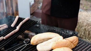 Siyah eldivenli bir aşçının ellerinin yakın çekimi, ızgarada sosisle rulo pişiren. Kömürle yemek pişirmek. Sokakta fast food.