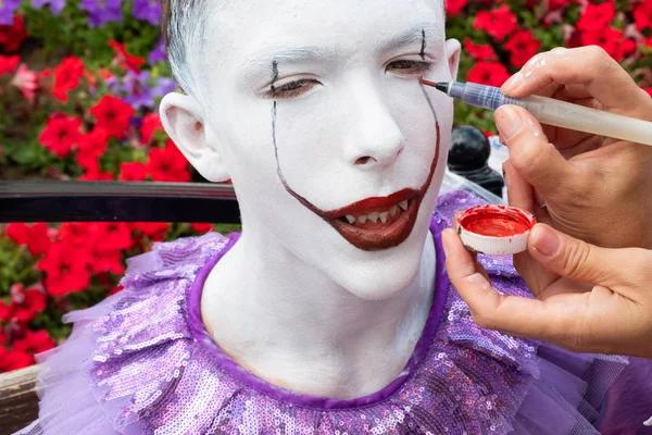 Kasachstan, kostanay, 19-08-16 street makeup. Das Gesicht eines Teenagers, dem die Hand eines Maskenbildners vor dem Hintergrund eines Blumenbeetes ein Bild eines Clowns oder Pantomimen zeichnet. Vorbereitung auf Karneval oder Halloween. — Stockfoto
