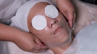 Beautician şeffaf bir film ile bir genç çocuğun yüzünü kapsar. Bir kadının ellerinin yakın çekim. Erkek müşteri güzellik salonunda yüz kozmetoloji prosedürü alır. Tıbbi akne temizliği.