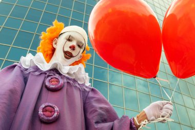 Bir gencin yüzünde sokak makyajı. Keskin dişleri ile bir palyaço görüntü bir çocuk elinde camdan yapılmış modern bir binanın arka planına karşı iki kırmızı balon tutar. Halloween.