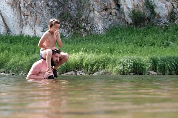 儿子坐在一位年轻父亲的肩上，双手牵着鼻子，然后跳入水中。在山河或湖中游泳。露营生活方式. — 图库照片
