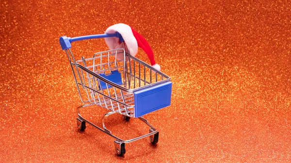 Santa Claus hat on a shopping trolley on a bright orange shiny background. The concept of Christmas shopping, New Year\'s discounts, online shopping, black Friday and a holiday sale. Copy space