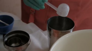 Shaker maskesi için bir karışım hazırlanması. Bir kadın toz döker ve metal bir bardağa su döker. Tıbbi, kimyasal veya kozmetik malzemeler ve mutfak eşyaları ile yeşil eldiven ellerin kapatılması.