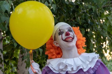Korkunç dişleri olan palyaço kostümlü bir adam neşeyle gülümsüyor. Elinde sarı balon var. Arka planda bir huş ağacının yeşil yaprakları. Karnaval ve Cadılar Bayramı kutlamaları cosplay.