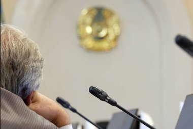 Gri saçlı bir yetkili eliyle başını destekler. Kazakistan haberleri için bulanık arka plan. Mikrofonlar, monitörler ve amblem. Devlet sorunları kavramı veya popüler sorunların çözümü.