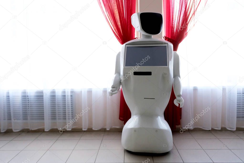 Information robot. A humanoid machine with manipulators instead of arms and a screen on the chest stands near a window with red curtains. Assistant and defender with artificial intelligence. Copyspace