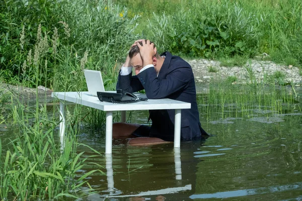 Problem, krise og rutine i erhvervslivet. En fyr i jakkesæt som forretningsmand, ekspedient eller leder i en vandpyt ved et hvidt bord med en bærbar computer, en telefon og holder fast i hovedet. Konceptkontor i sumpen . - Stock-foto