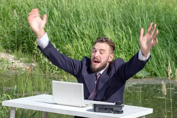 Ufficio nella palude. Un uomo d'affari con la barba si siede a un tavolo bianco con un computer portatile e un telefono e grida aiuto. Movimento a mano con grasso. Concetto di problemi, crisi o routine nelle imprese . — Foto Stock