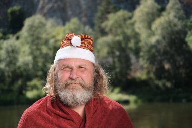Noel Baba şapkalı ve kırmızı ceketli yaşlı, karizmatik bir adamın portresi. Yaz manzarası ormanı. Sıcak ülkelerde Noel ve Yeni Yıl tatili ya da tatil kavramı.
