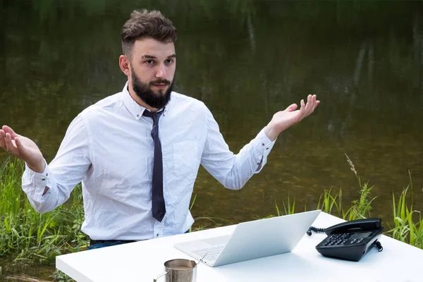 A businessman or an office worker with a beard throws up his hands to the side from hopelessness and routine work. Table with a laptop and a phone on the river. Freelancer concept or dream of vacation