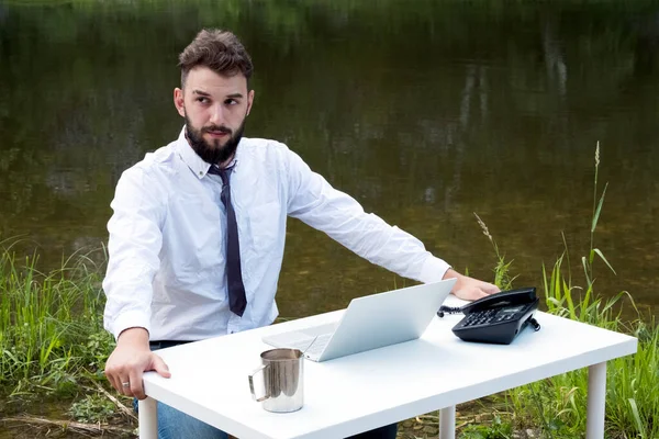 A tired businessman or office worker with a beard sits at a table on the nature with a laptop and a phone on the background of a river and reeds and thinks looking to the side. Concept of freelance.