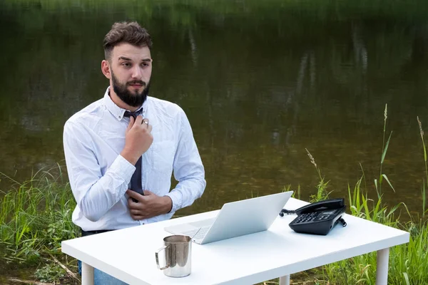 Morning businessman in a white shirt. A guy with a beard sits at a table with a laptop, a mug of coffee against of the river and straightens his tie. The concept of routine and remote work.