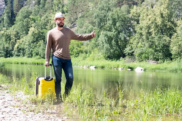 El viajero en el sombrero levantó la mano y levantó el pulgar como si estuviera cogiendo un taxi. Triste joven hipster haciendo autostop a lo largo de un río de montaña con una maleta amarilla — Foto de Stock