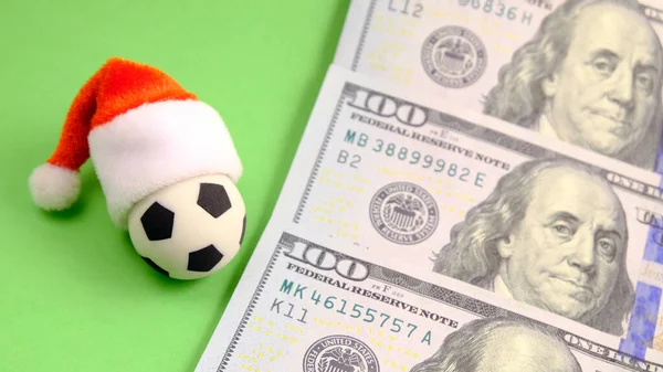 Souvenir soccer ball in a red Santa Claus hat next to three hundred US dollars on a green background. Christmas sporting events or New Year sports bets.