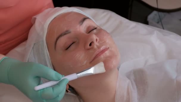 As mãos de um cosmetologista usam uma escova para aplicar gel na pele de uma linda garota europeia. Um tratamento rejuvenescedor e relaxante no centro de beleza . — Vídeo de Stock