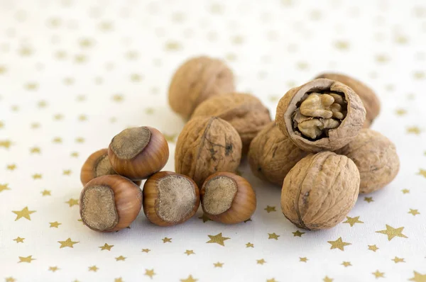 Walnuts and hazelnuts in hard shells, pile on white Christmas tablecloth with golden stars, one walnut with broken shell