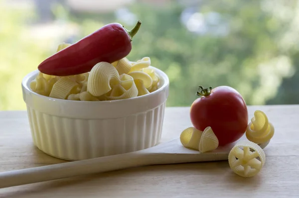 Various mix of pasta in white baking bowl on wooden table and ripened red jalapeno pepper, wooden spoon and red tomato