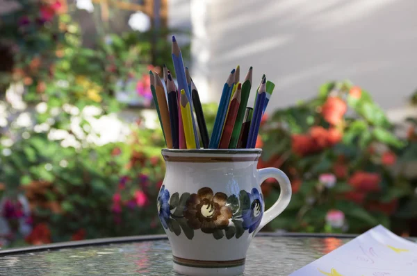 Stack of color pencils in the flower mug on the glass table, outdoors, greenery around, sunny