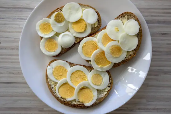 Skivade hårdkokta ägg på vanligt tjeckiskt bröd med smör på vit tallrik på ljust träbord — Stockfoto