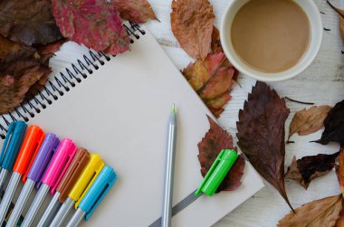 Sonbahar kavramı. Sonbahar yaprakları, kahve fincanı, defter ve renkli kalemler üzerinde ahşap masa vardır. Üstten görünüm,