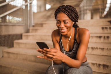 Merdivenlerde oturan ve Smartphone cep telefonu ile en sevdiğiniz müzik dinlerken Afrikalı-Amerikalı kadın gülümsedi