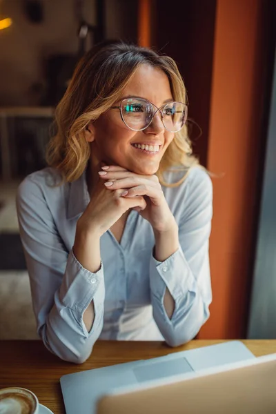 Beautiful young woman with glasses is sitting in cafe and looking through a window in a distance