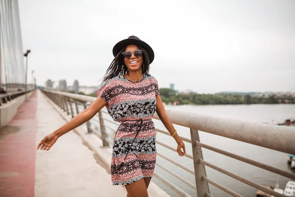 Smiling black woman with hat is walking over a bridge and enjoy her free time