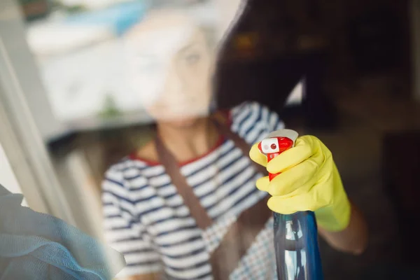 Young woman wearing apron and gloves and using sprayer to clean windows at her home