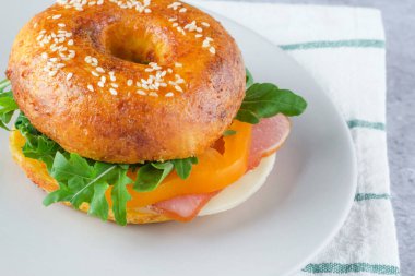 Jambon, peynir ve taze yeşilliklerle dolu lezzetli bir susamlı sandviç hafif bir tabakta oturur. Yemek desenli bir yüzeyde hazırlanır, tazeliğini ve rengini vurgular. Yüksek kalite fotoğraf