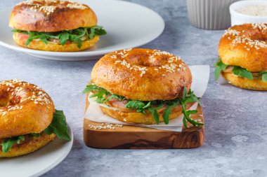 Yeşillik ve jambonla dolu, ahşap bir tahta ve tabakta üç taze pişmiş simit. Bagel kahvaltı sandviçi eşsiz bir lezzet katar. Yüksek kalite fotoğraf