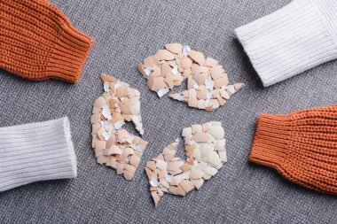 Gri desenli bir yüzeyde, örgülü kollarla çevrili kırık yumurta kabuğu parçalarıyla yaratıcı geri dönüşüm sembolü. Tekstil endüstrisinde yumurta kabukları sürdürülebilir bir malzeme olarak kullanılıyor. Organik tekstil yeniliği. Üst Manzara. Yüksek kalite fotoğraf