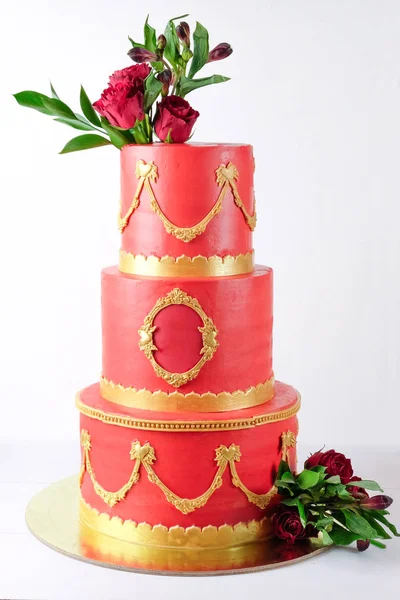 Three-tiered red wedding cream cake decorated with flowers and gold elements on a white wooden table. Picture for a menu or a confectionery catalog.