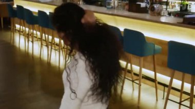 Kıvırcık saçlı güzel kız barda dans ediyor. Mutlu sevimli esmer beyaz kazaklı sahiptir boş bir kafeye çevrili